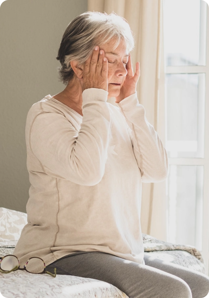 Older woman sitting on a bed by a sunlit window, pressing her temples in apparent headache.