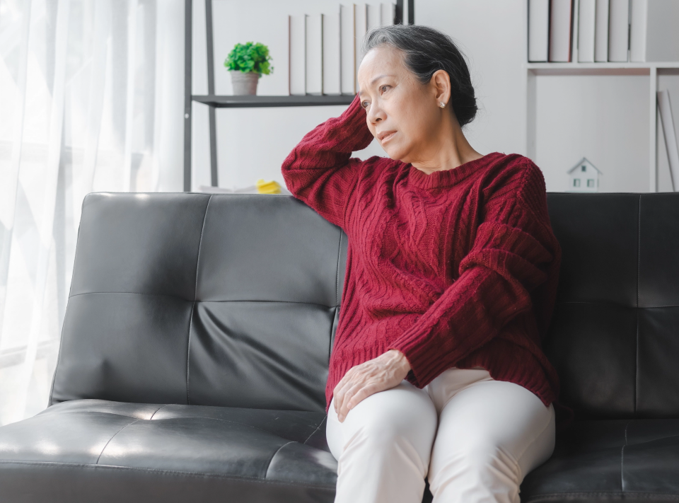 Older woman sitting on a black leather couch, resting her head on her hand and looking contemplative in a bright living room.