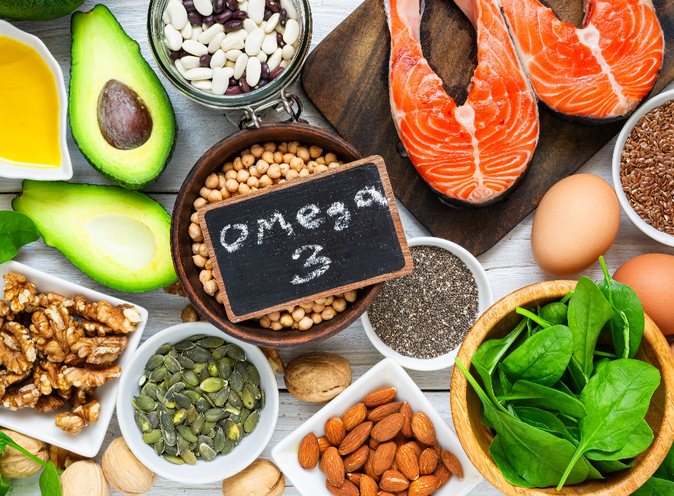 Omega-3 rich foods on a board: salmon fillets, avocado halves, nuts, seeds, greens, eggs, with a chalkboard reading Omega-3.