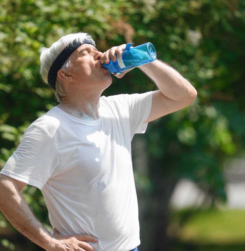 Man in a white t-shirt wearing a black headband drinks from a blue water bottle outside, with greenery in the background.