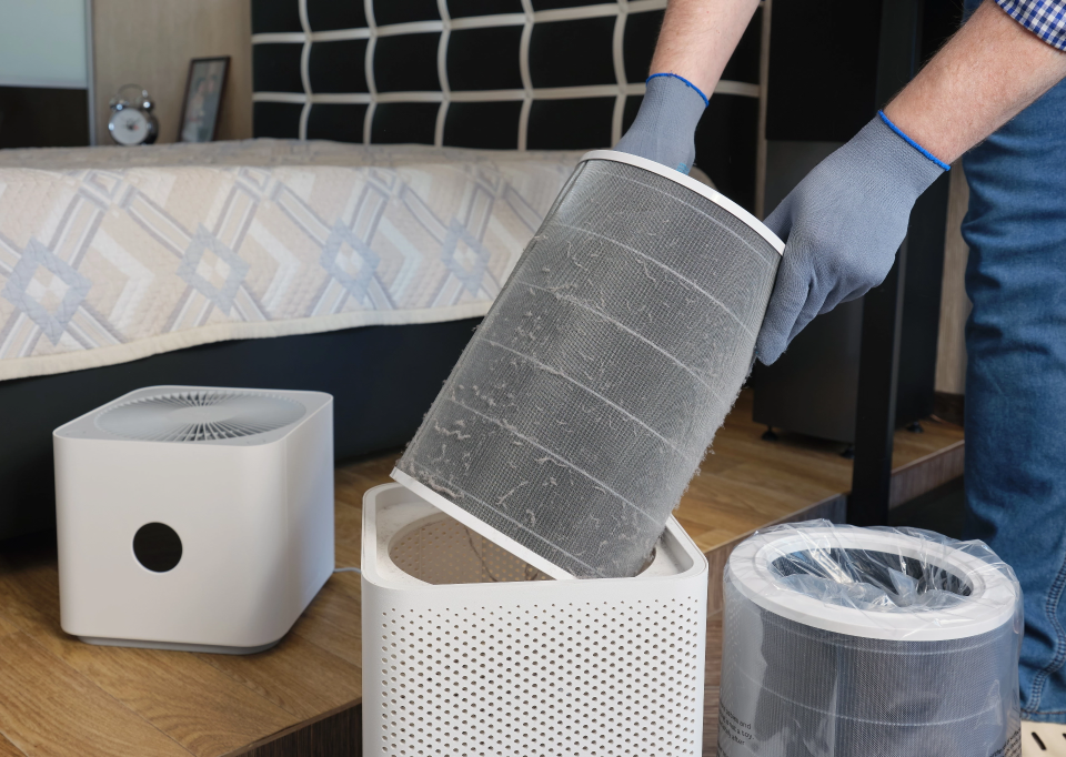 Person wearing gloves inserts a cylindrical filter into a white air purifier unit on a wooden floor.