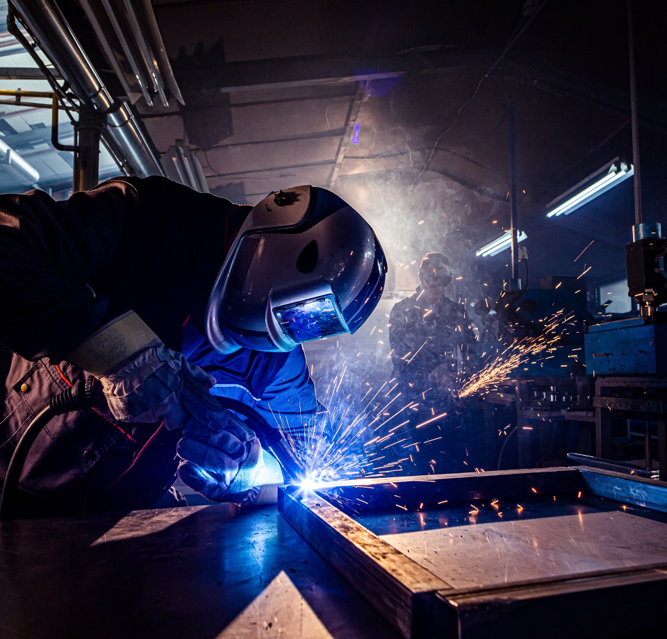 Welder wearing helmet and gloves, welding metal with a bright arc and sparks in a workshop.
