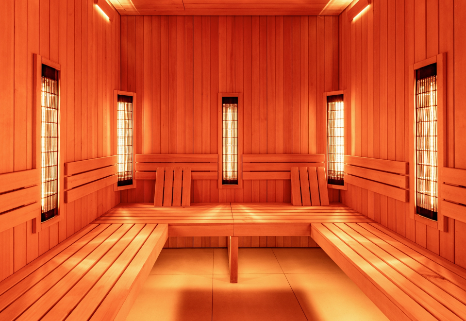 Wooden sauna room with long benches along three walls and tall infrared heaters emitting warm light.