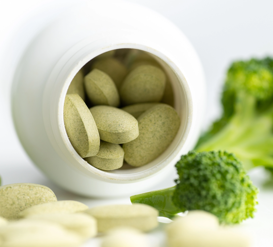 Green tablets spilling from a white bottle beside broccoli florets, suggesting a dietary supplement.
